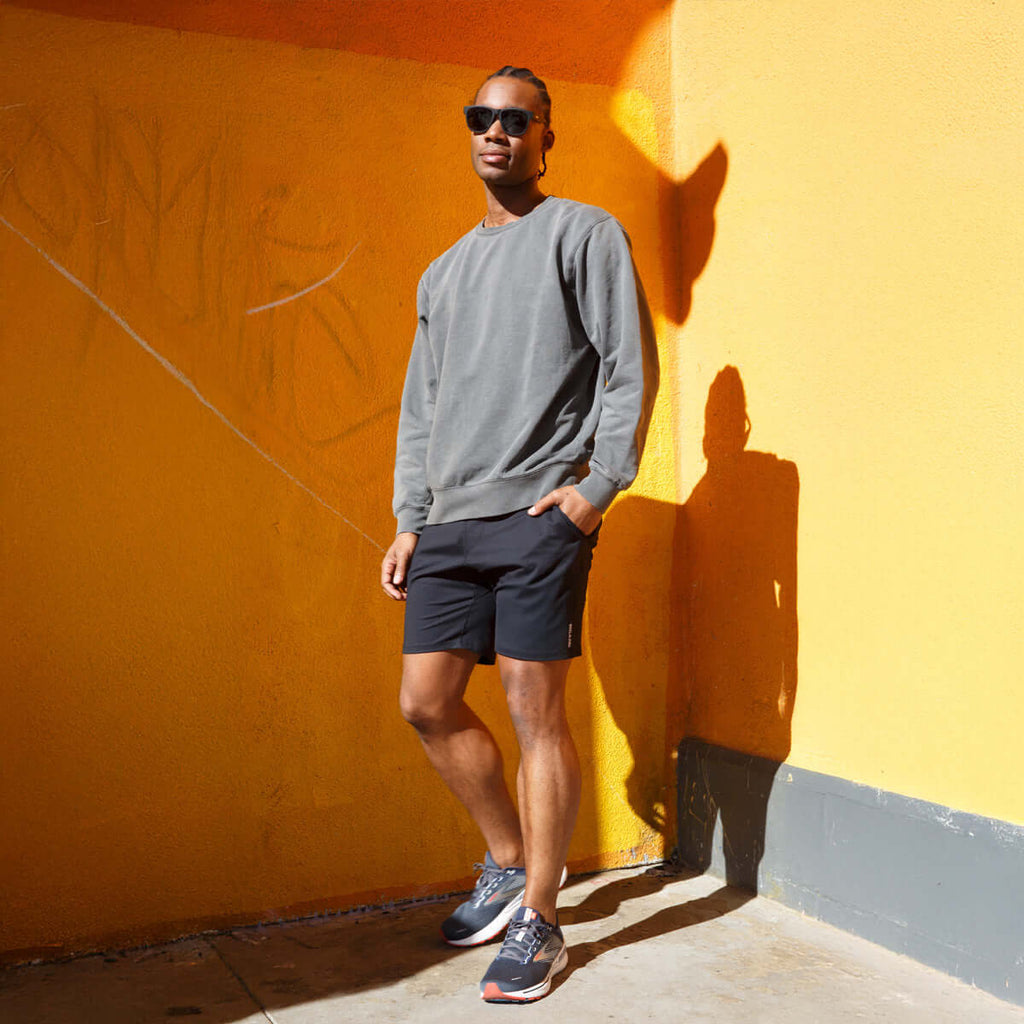 Model wearing 4-way stretch swerve shorts in Crow Black and a gray sweatshirt, standing against a colorful wall.