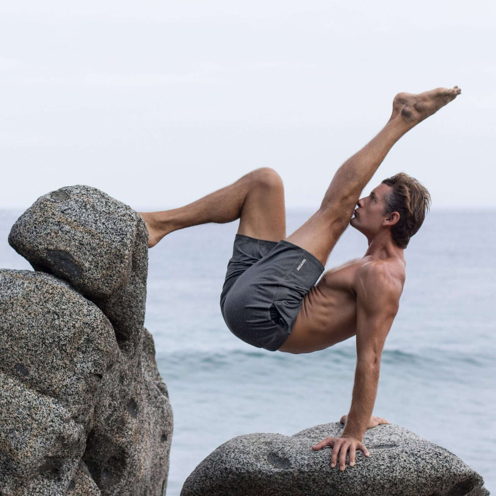 Man performing a yoga pose on rocks by the ocean while wearing 4-way stretch swerve shorts in heather grey.